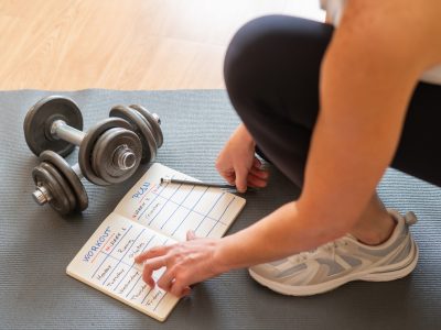 Female Athlete Organizing Fitness Plan On Notepad While Sitting On Yoga Mat With Dumbbells, Creating Healthy Daily And Weekly Exercise Routines, Promoting Active Lifestyle Goals, Morning Motivation