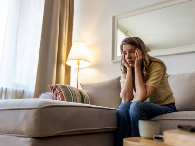 A woman sits on a beige sofa in a bright living room, hands on her face, looking worried and tired. Soft lamp light, cushions, and a calm environment contrast with her stressed mood.